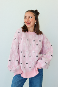 A smiling woman with her hair in two buns wears a light pink fuzzy knit sweater with a repeating black and white ghost pattern, paired with blue jeans, against a plain light gray background.