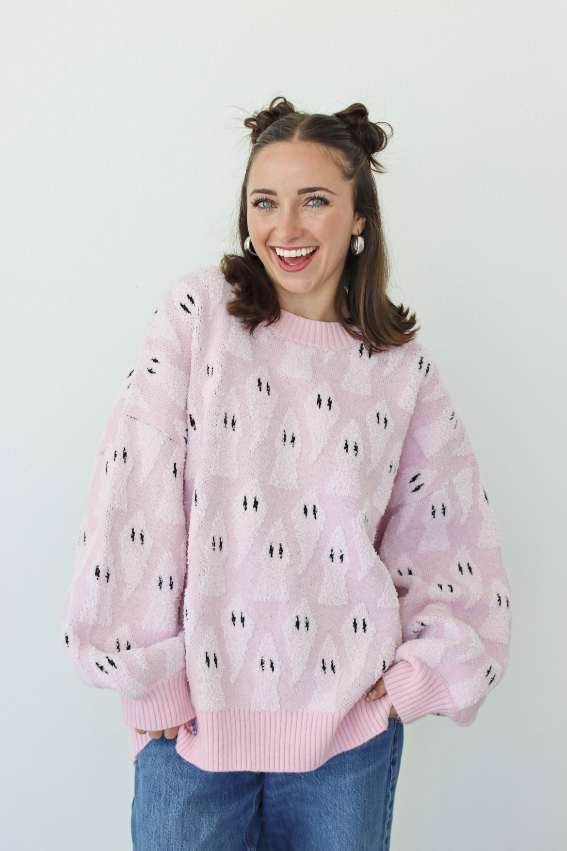 A smiling woman with her hair in two buns wears a light pink fuzzy knit sweater with a repeating black and white ghost pattern, paired with blue jeans, against a plain light gray background.