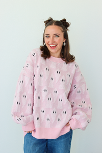 A smiling woman with her hair in two buns wears a light pink fuzzy knit sweater with a repeating black and white ghost pattern, paired with blue jeans, against a plain light gray background.
