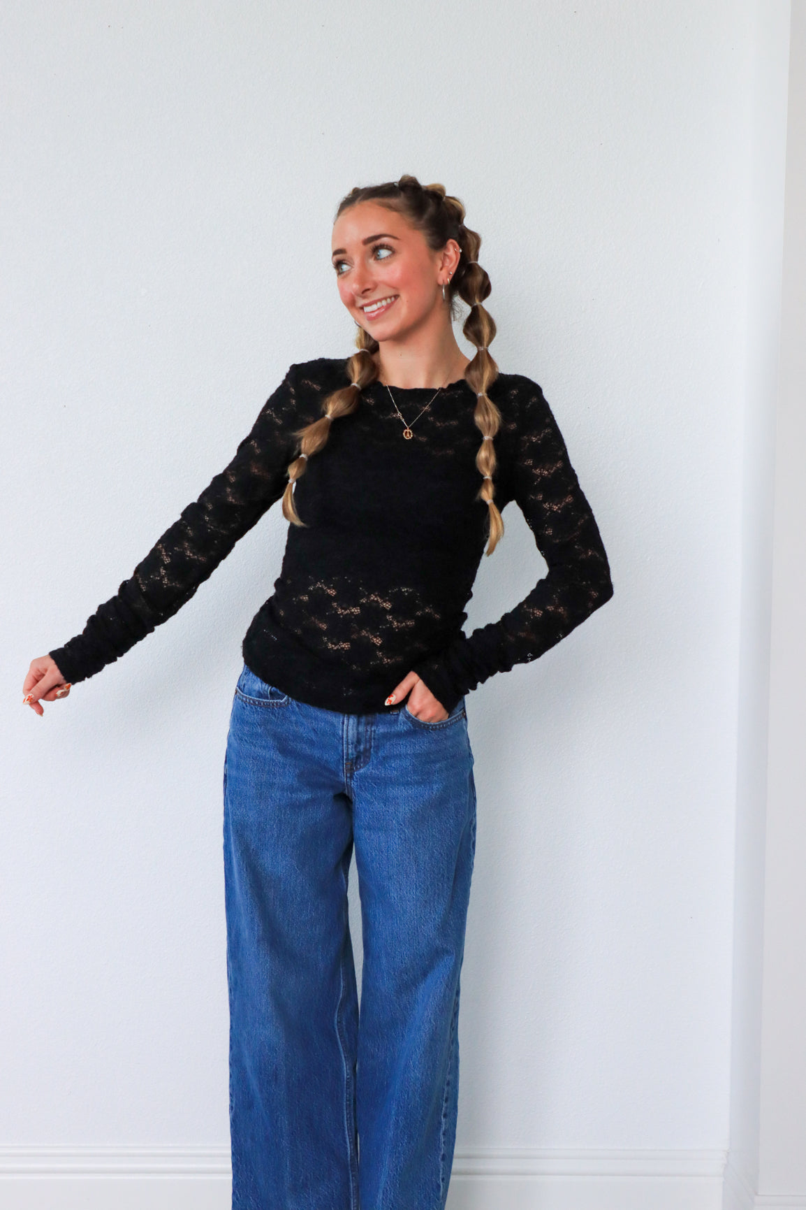 Woman wearing a black lace top and blue jeans against a white wall