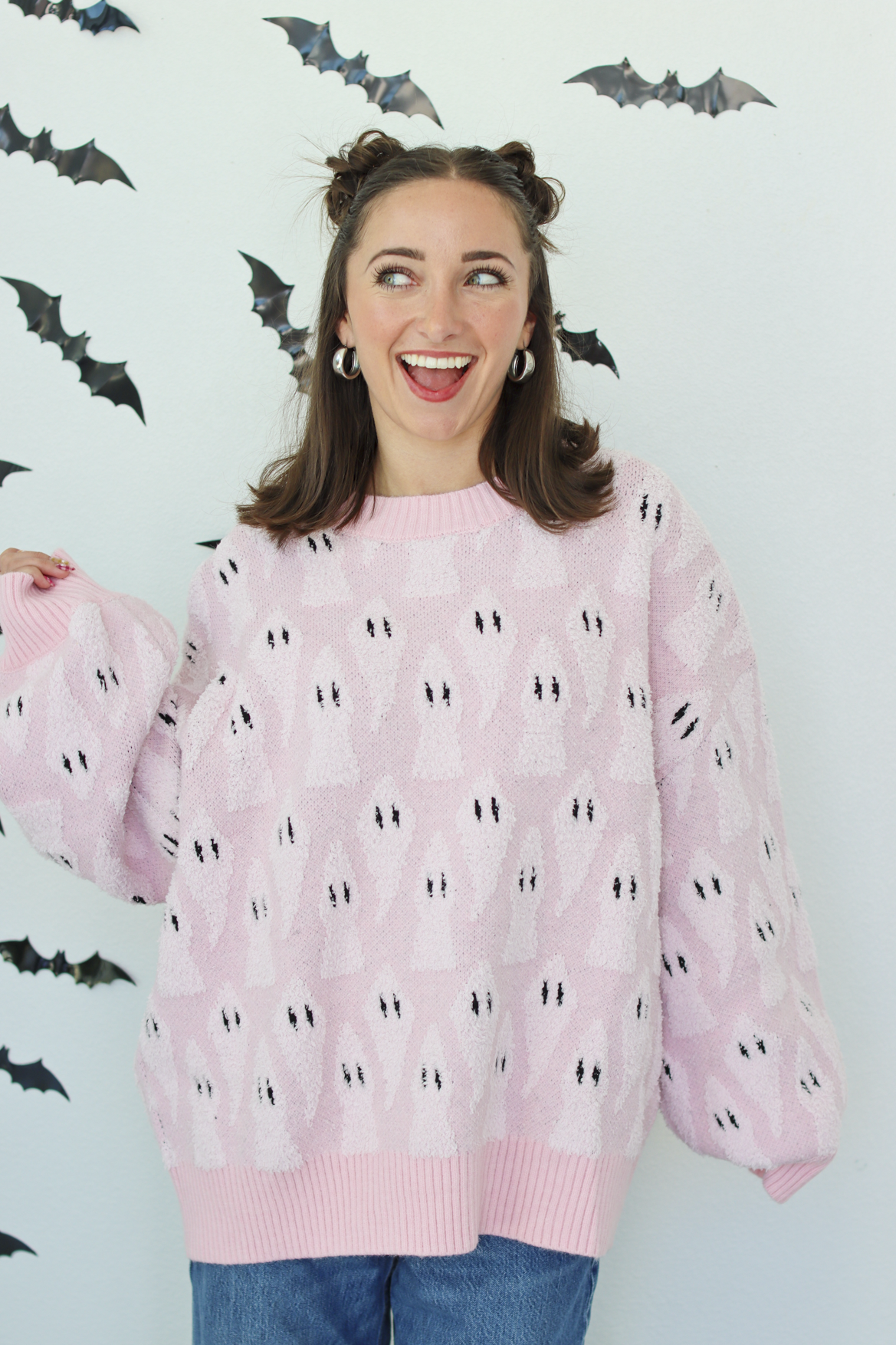 A woman wearing a light pink knit sweater with a repeating black and white ghost pattern, posed against a white wall decorated with black bat cutouts.