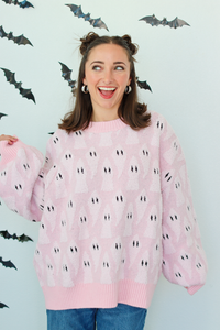 A woman wearing a light pink knit sweater with a repeating black and white ghost pattern, posed against a white wall decorated with black bat cutouts.