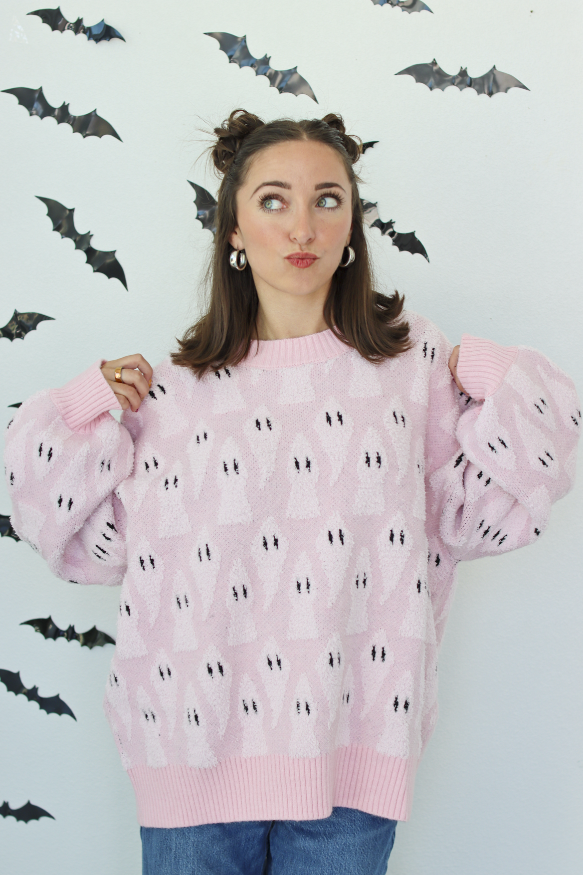A woman wearing a light pink knit sweater with a repeating black and white ghost pattern, posed against a white wall decorated with black bat cutouts.