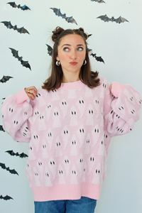 A woman wearing a light pink knit sweater with a repeating black and white ghost pattern, posed against a white wall decorated with black bat cutouts.