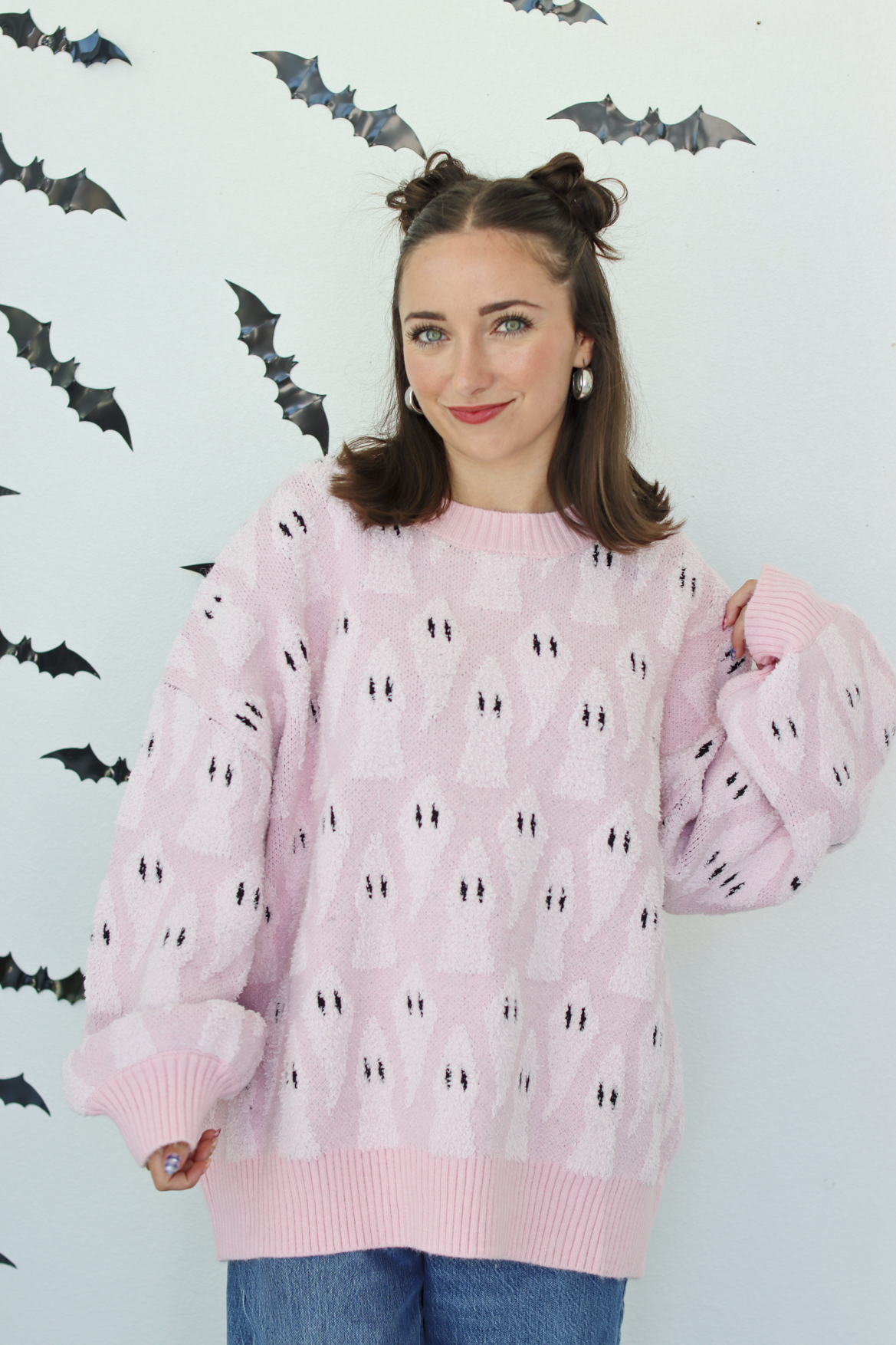 A woman wearing a light pink knit sweater with a repeating black and white ghost pattern, posed against a white wall decorated with black bat cutouts.