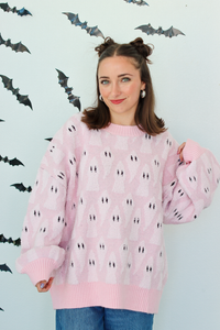 A woman wearing a light pink knit sweater with a repeating black and white ghost pattern, posed against a white wall decorated with black bat cutouts.