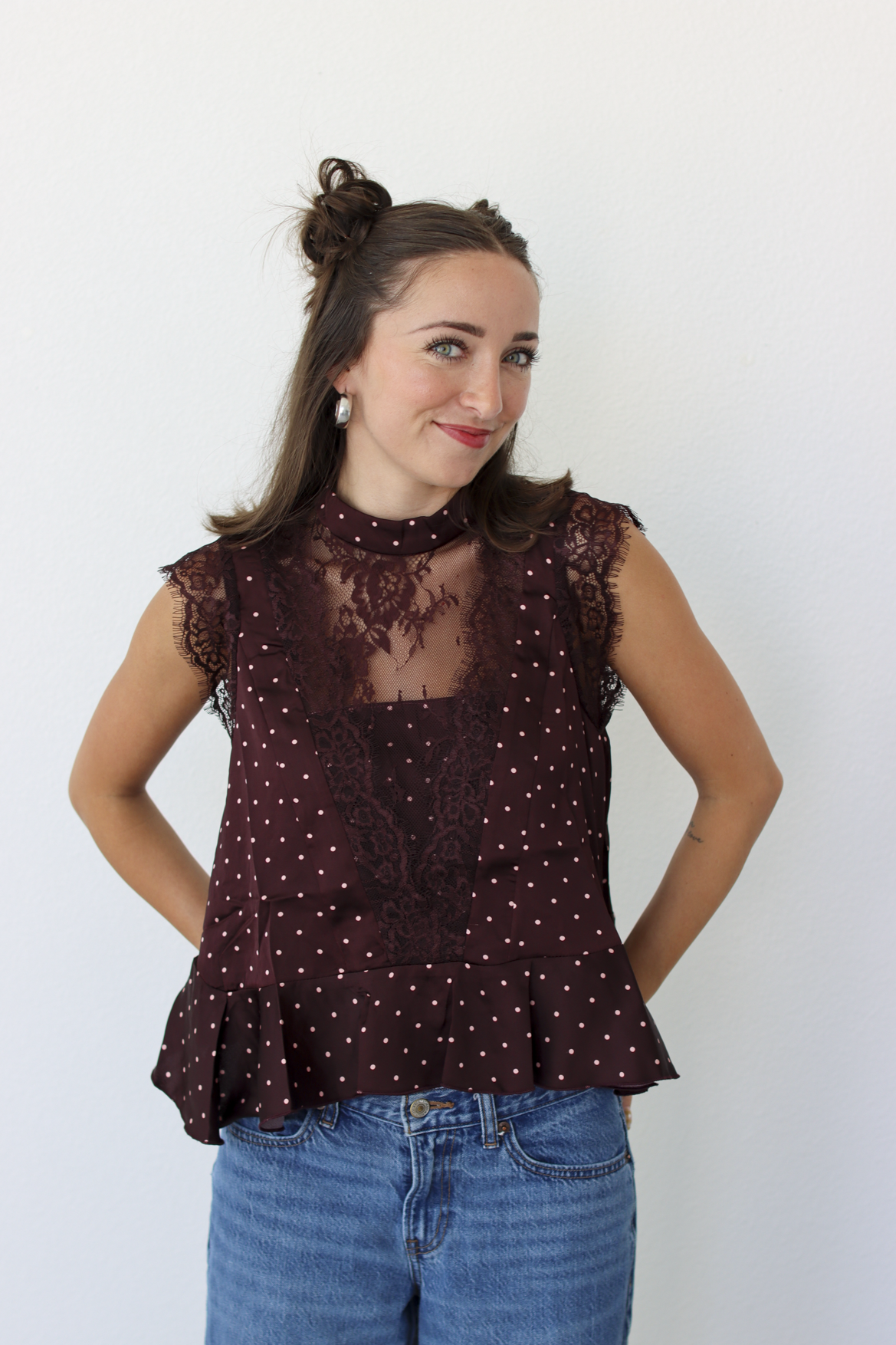 A woman modeling a sleeveless burgundy top with white polka dots and a sheer lace yoke, paired with blue jeans, against a plain white background.