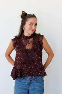 A woman modeling a sleeveless burgundy top with white polka dots and a sheer lace yoke, paired with blue jeans, against a plain white background.