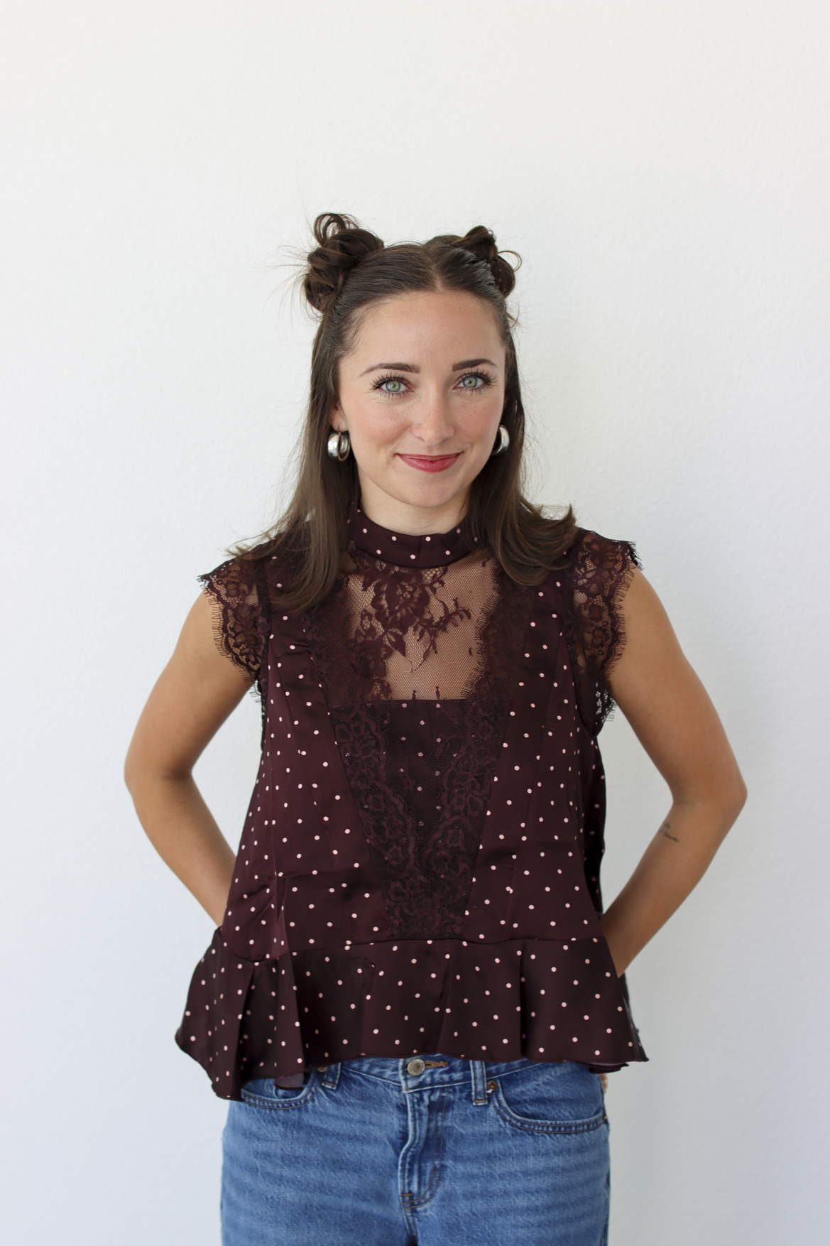 A woman modeling a sleeveless burgundy top with white polka dots and a sheer lace yoke, paired with blue jeans, against a plain white background.