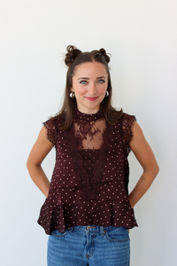 A woman modeling a sleeveless burgundy top with white polka dots and a sheer lace yoke, paired with blue jeans, against a plain white background.