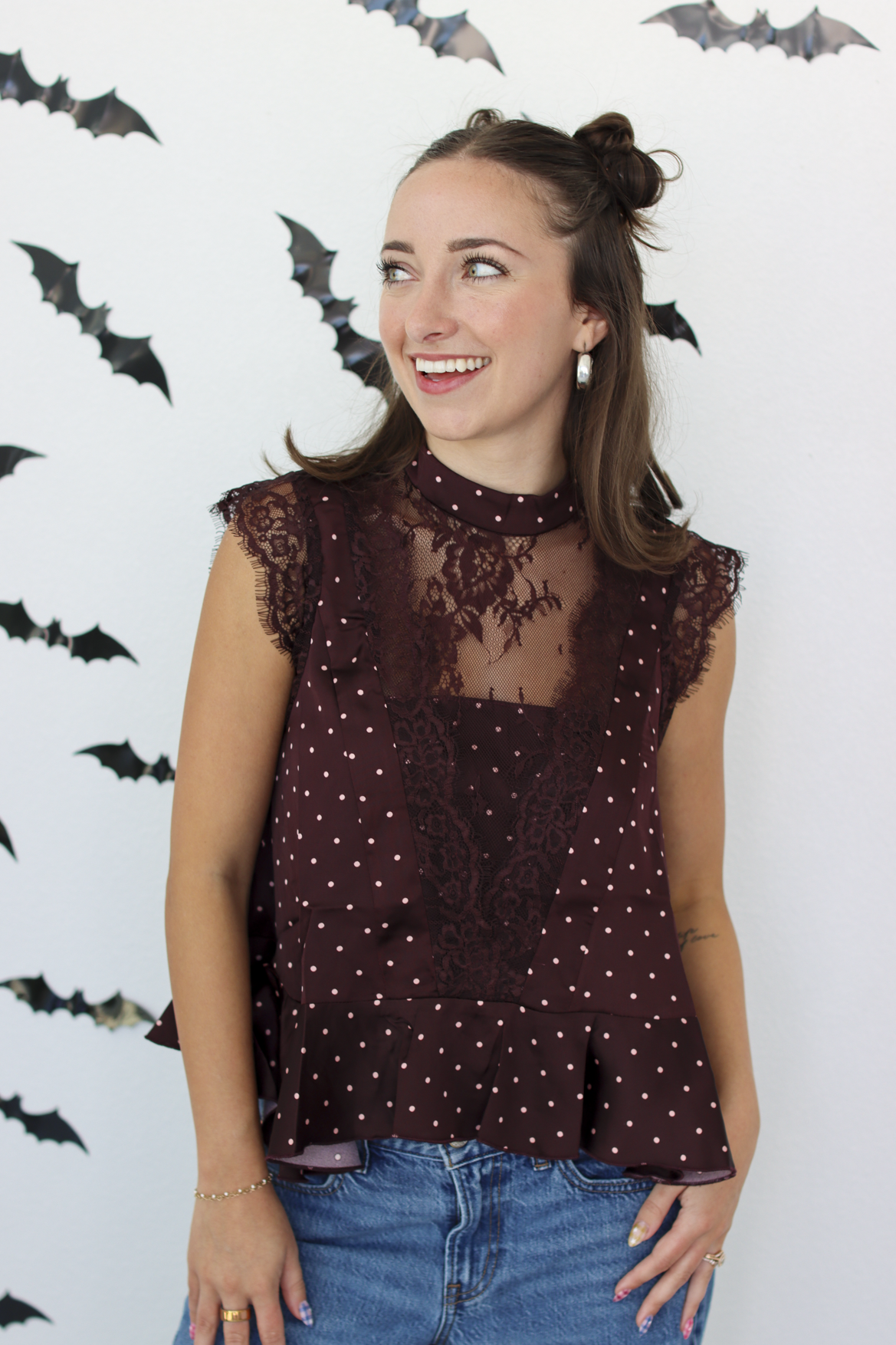 A woman wearing a sleeveless burgundy top with white polka dots and lace detailing, paired with blue jeans, standing against a white wall decorated with black bat cutouts.