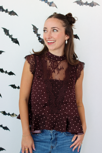 A woman wearing a sleeveless burgundy top with white polka dots and lace detailing, paired with blue jeans, standing against a white wall decorated with black bat cutouts.