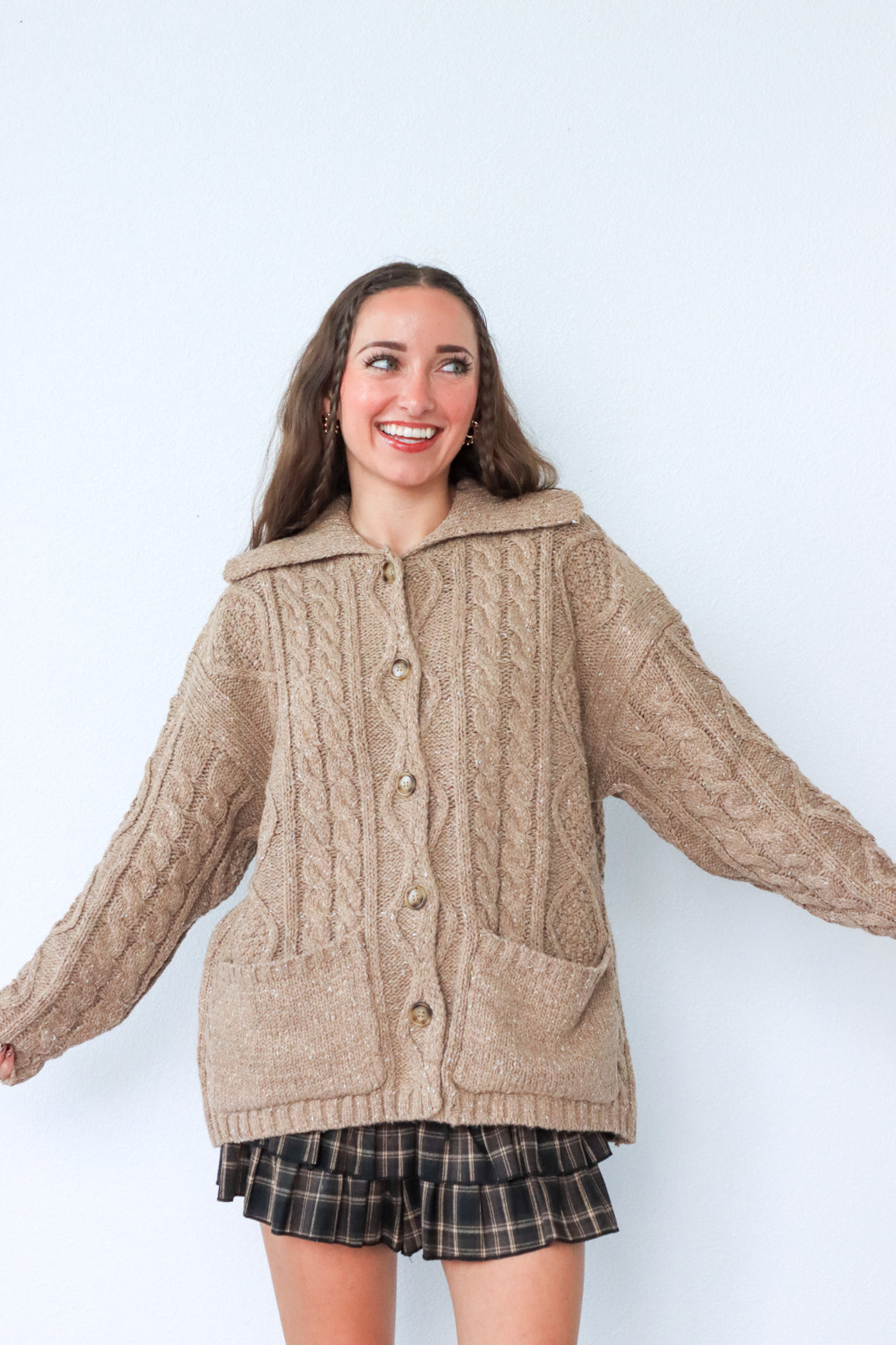 Woman wearing a beige knitted cardigan with a plaid skirt against a white background