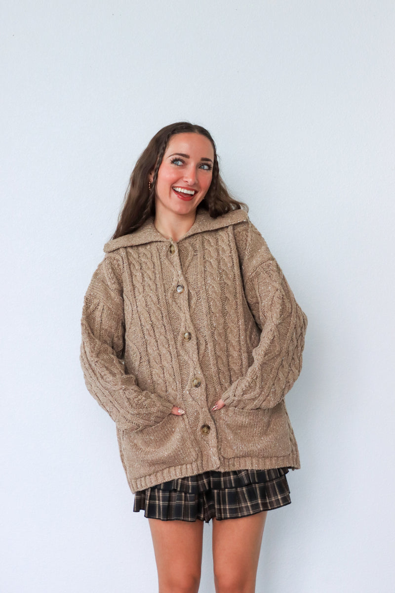 Person wearing a beige cable knit cardigan against a white background
