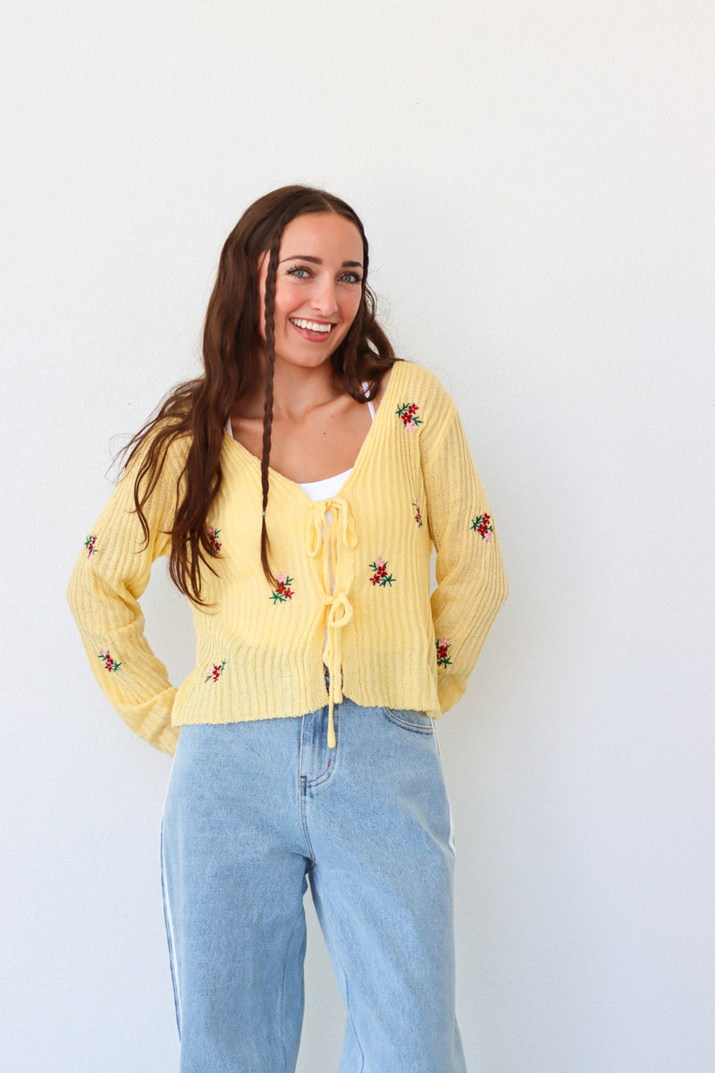 Woman wearing a yellow embroidered cardigan and light blue jeans against a white background