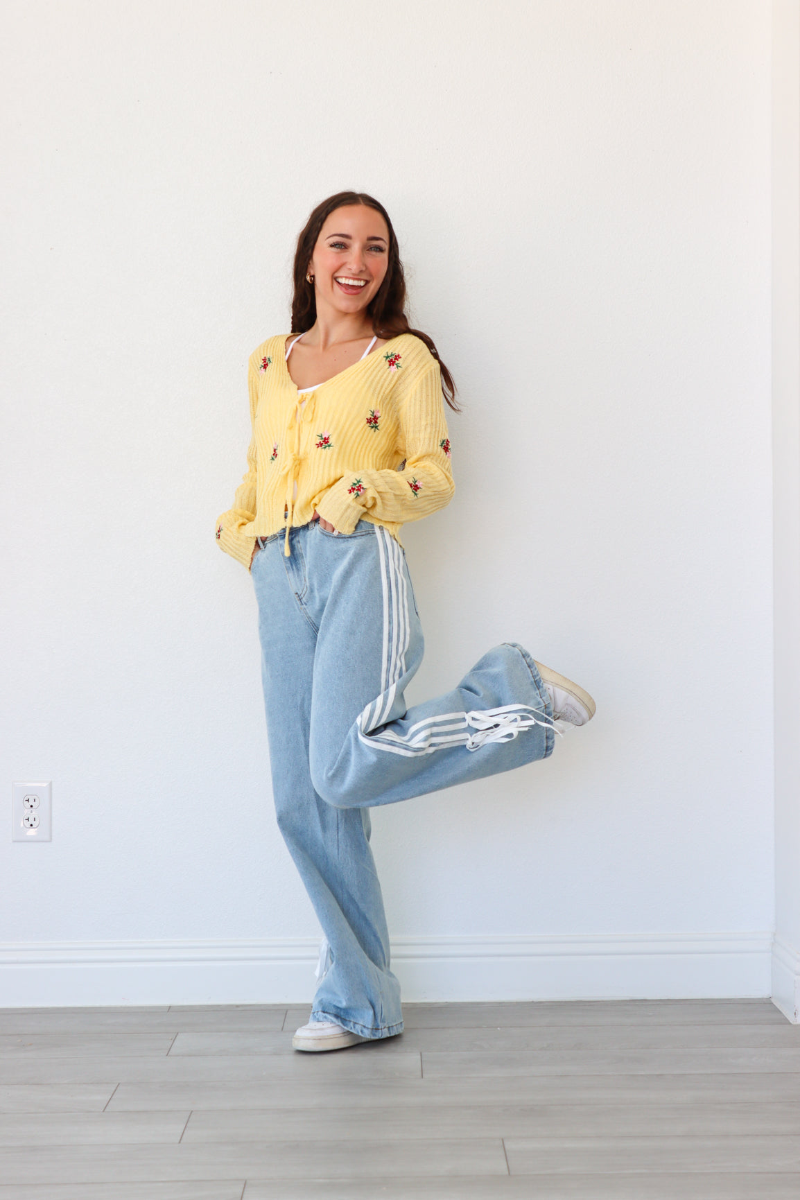Woman wearing a yellow sweater and light blue pants with white stripes, standing against a white wall.
