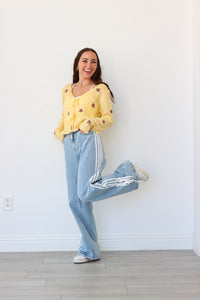 Woman wearing a yellow sweater and light blue pants with white stripes, standing against a white wall.