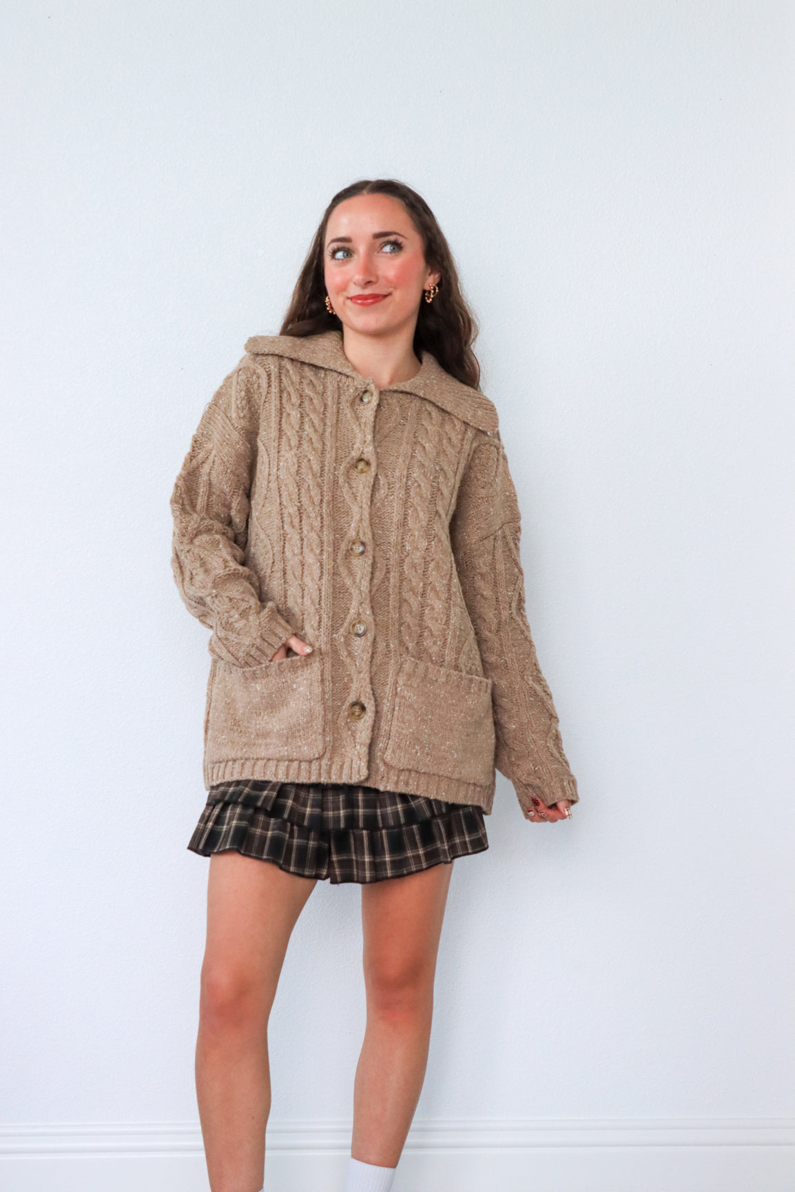 Woman wearing a beige cardigan over a plaid skirt against a white background