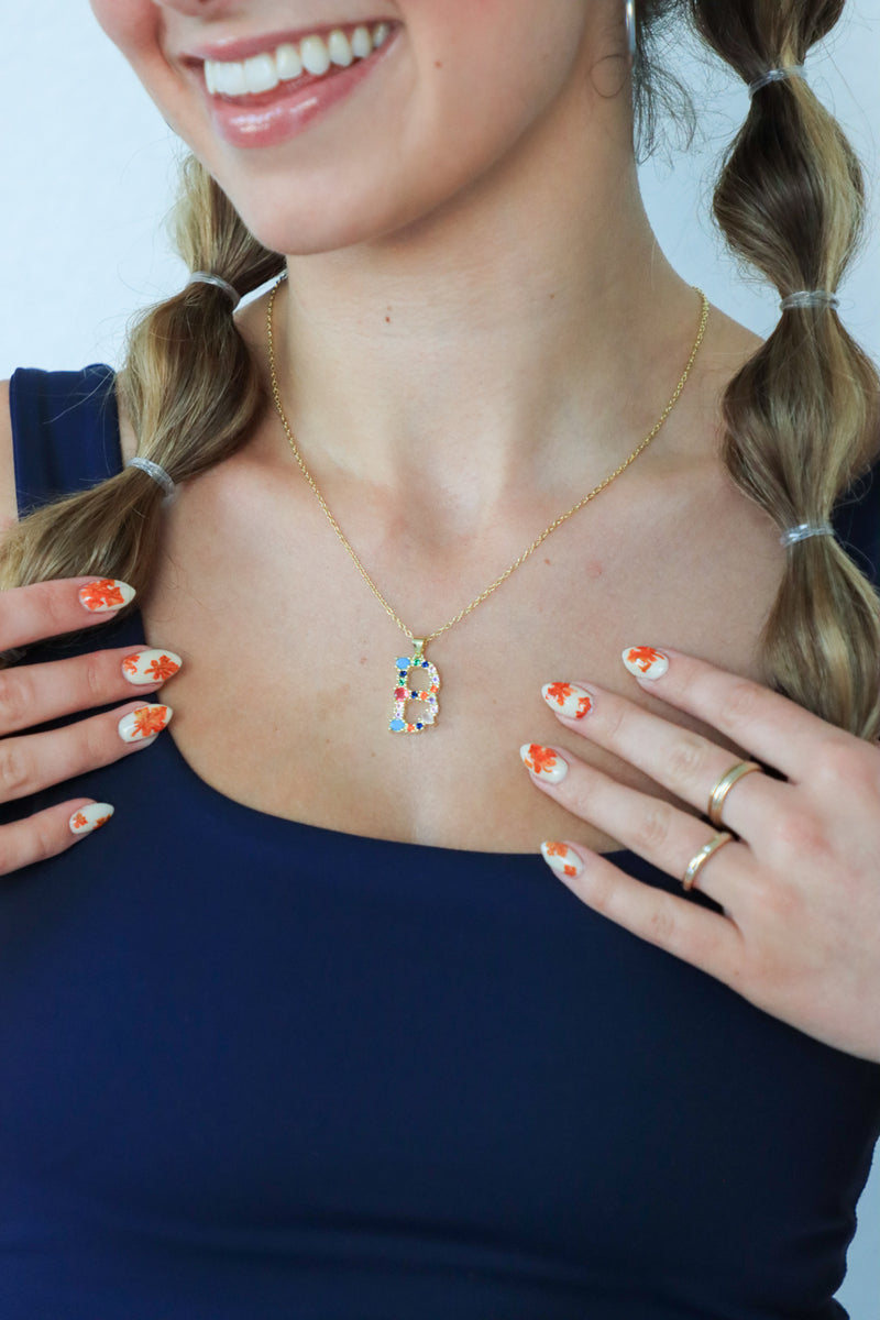 Woman wearing a gold necklace with a colorful pendant, smiling against a light blue background.