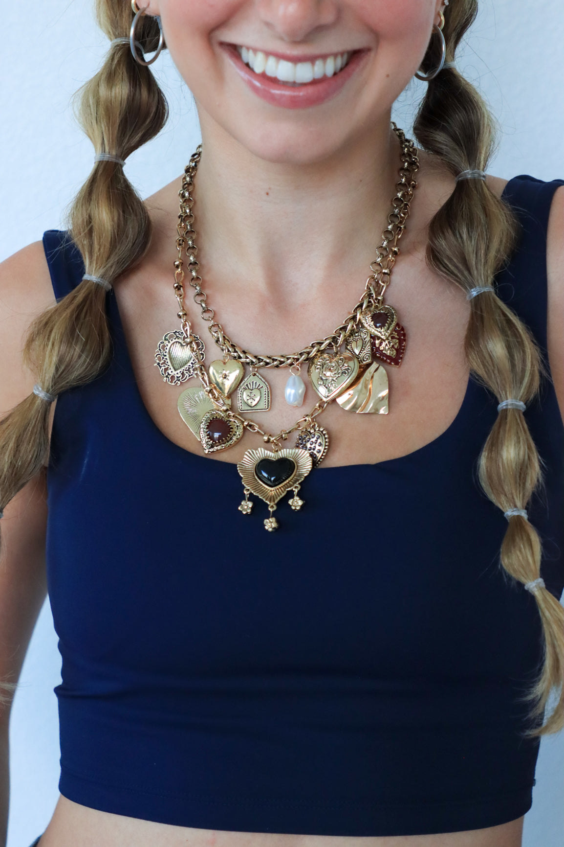 Woman wearing a large, ornate necklace with heart and circular pendants against a plain background.