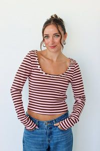 A woman wearing a long-sleeve top with horizontal pink and brown stripes, featuring decorative buttons around the scoop neckline, paired with blue jeans against a plain white background.