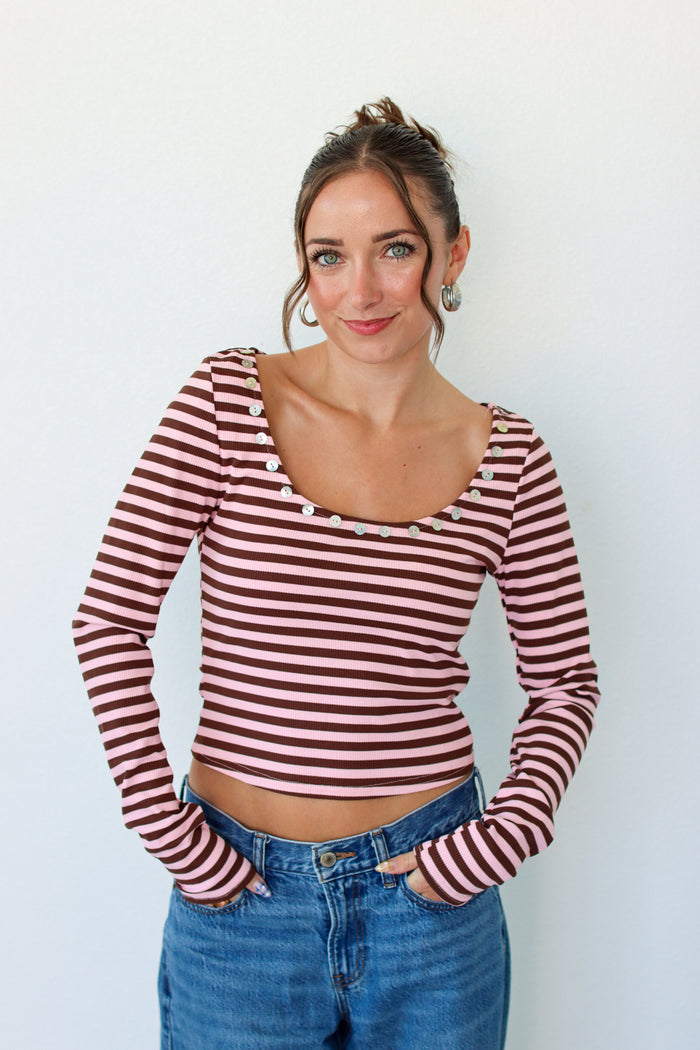 A woman wearing a long-sleeve top with horizontal pink and brown stripes, featuring decorative buttons around the scoop neckline, paired with blue jeans against a plain white background.