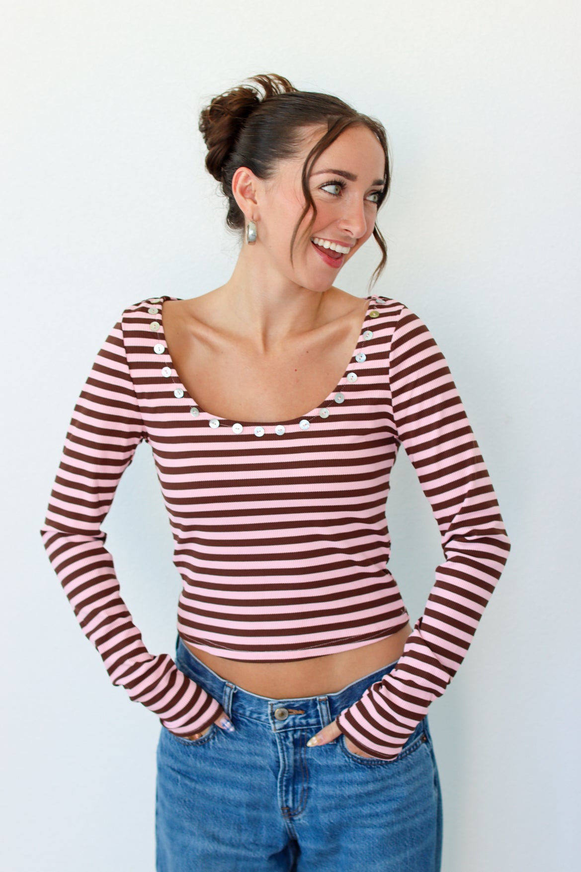 A woman wearing a long-sleeve top with horizontal pink and brown stripes, featuring decorative buttons around the scoop neckline, paired with blue jeans against a plain white background.