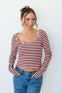 A woman wearing a long-sleeve top with horizontal pink and brown stripes, featuring decorative buttons around the scoop neckline, paired with blue jeans against a plain white background.