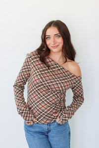 A woman with long brown hair wearing a brown and beige plaid off-the-shoulder long-sleeve top with red lines, paired with blue jeans, standing against a plain white wall.