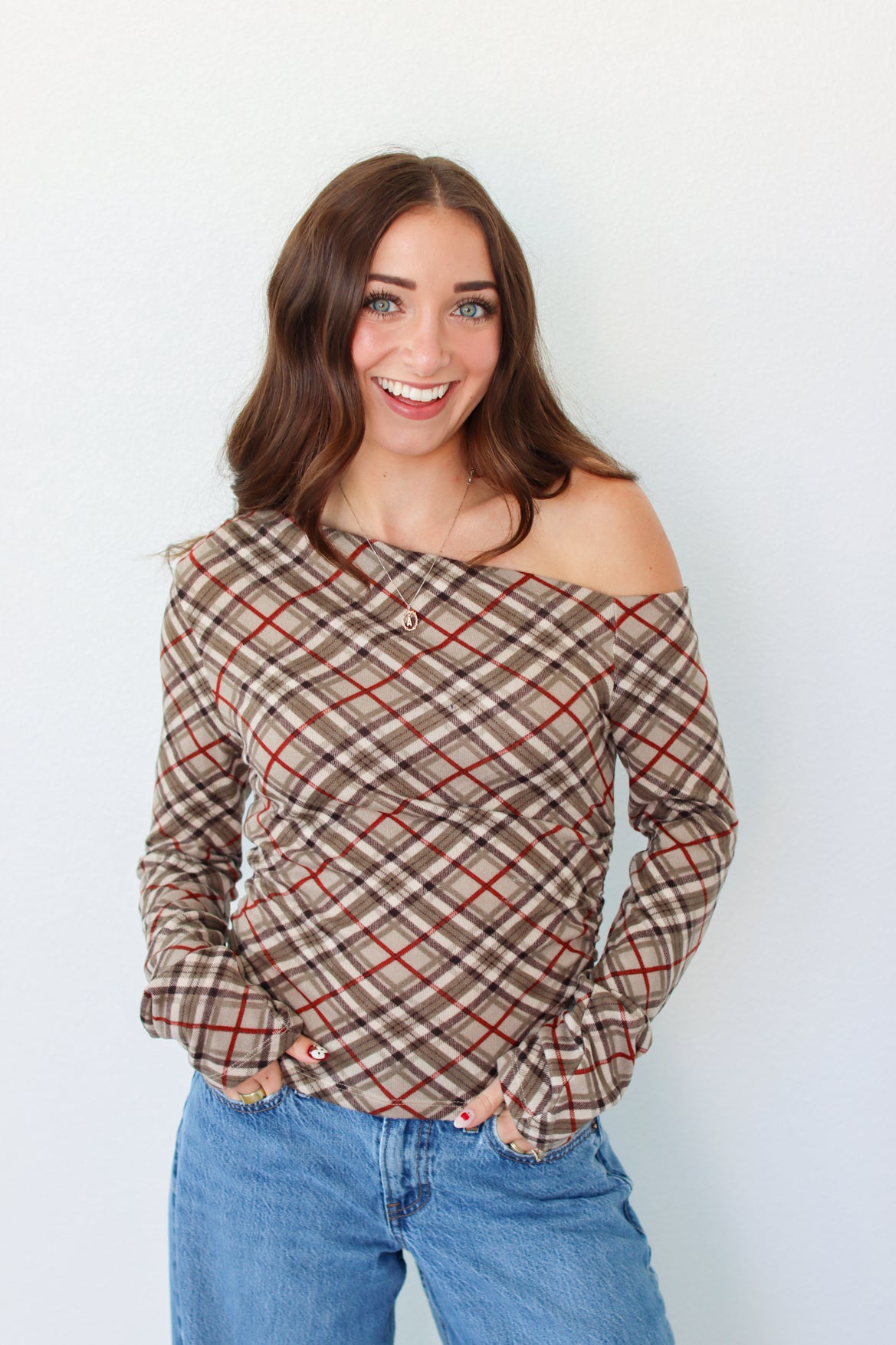 A woman with long brown hair wearing a brown and beige plaid off-the-shoulder long-sleeve top with red lines, paired with blue jeans, standing against a plain white wall.