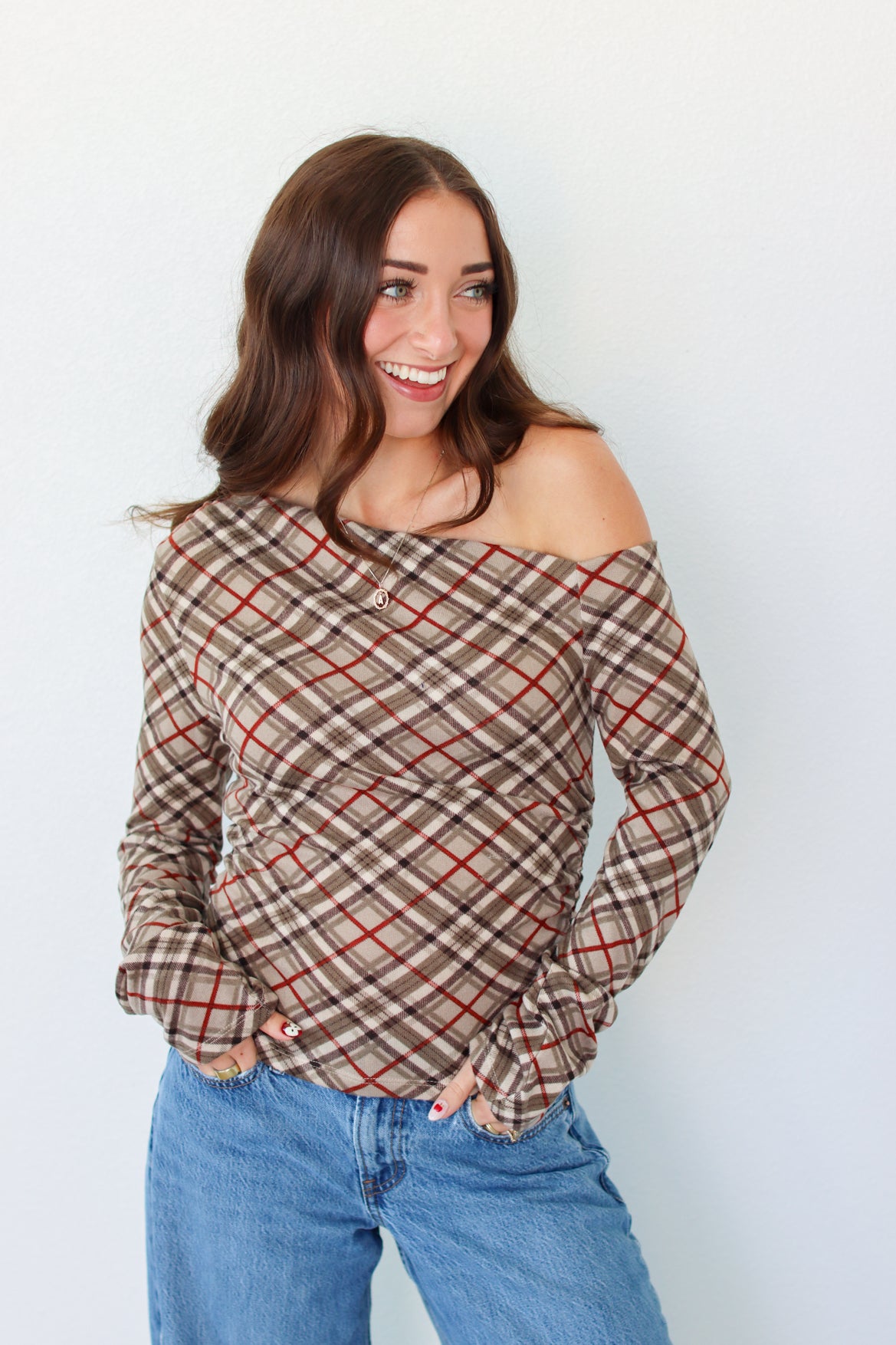 A woman with long brown hair wearing a brown and beige plaid off-the-shoulder long-sleeve top with red lines, paired with blue jeans, standing against a plain white wall.