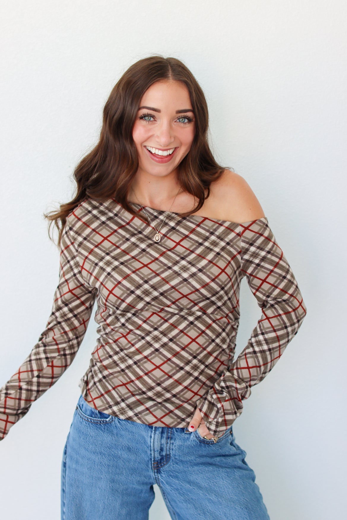 A woman with long brown hair wearing a brown and beige plaid off-the-shoulder long-sleeve top with red lines, paired with blue jeans, standing against a plain white wall.