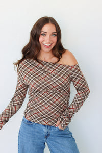 A woman with long brown hair wearing a brown and beige plaid off-the-shoulder long-sleeve top with red lines, paired with blue jeans, standing against a plain white wall.