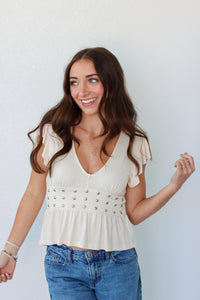 A woman with long brown hair smiling and looking to the side, wearing a cream-colored short-sleeve peplum top with silver studs and blue jeans, standing against a light gray wall.