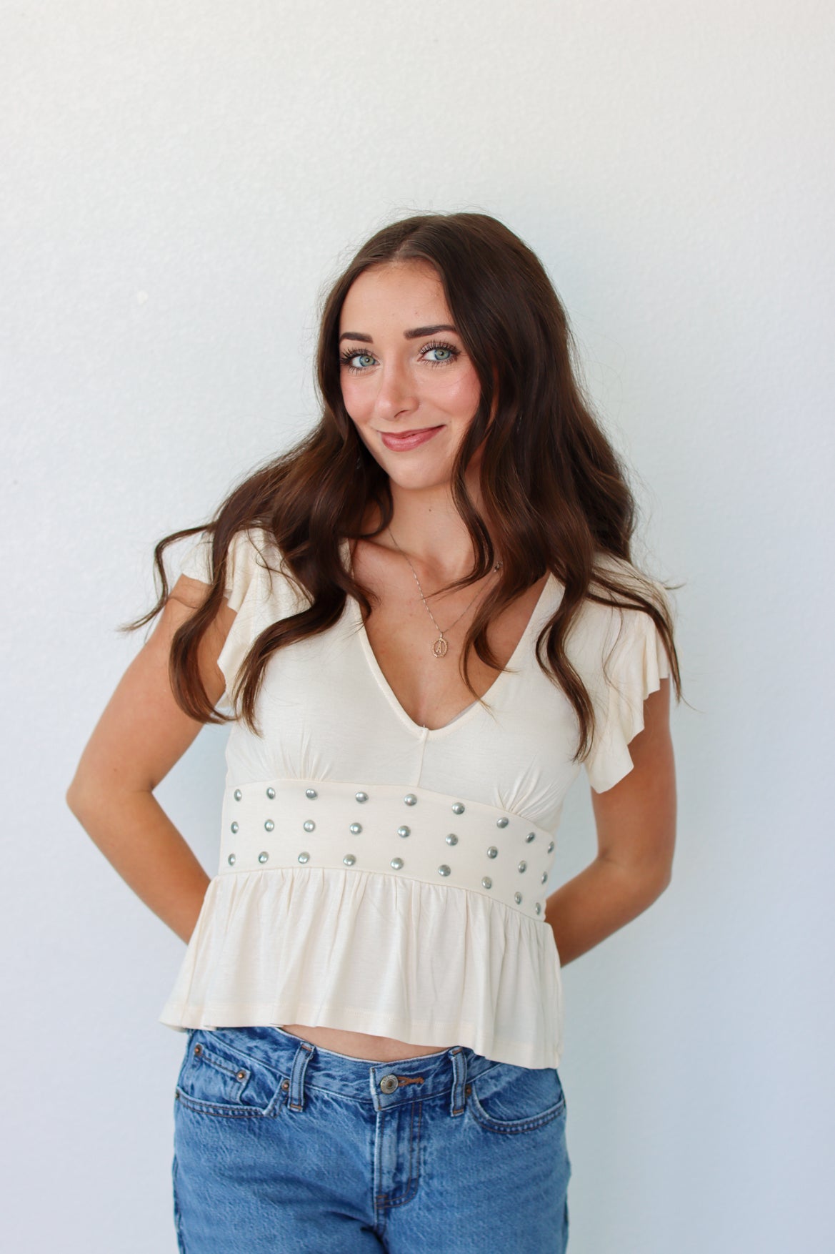 A woman with long brown hair wearing a cream-colored ruffle-sleeve crop top with silver stud embellishments and blue jeans, standing against a plain white wall.