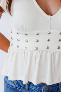 Close-up of a person wearing a cream-colored sleeveless top with a ruffled hem and a wide band of silver studs, paired with blue jeans.