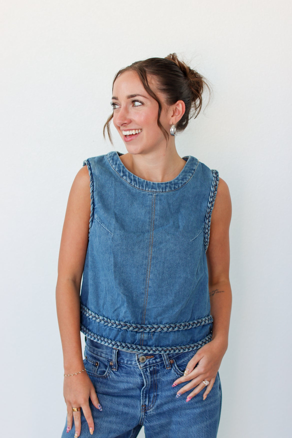 A woman wearing a sleeveless blue denim vest with braided trim details, paired with blue jeans, posing against a plain white wall.