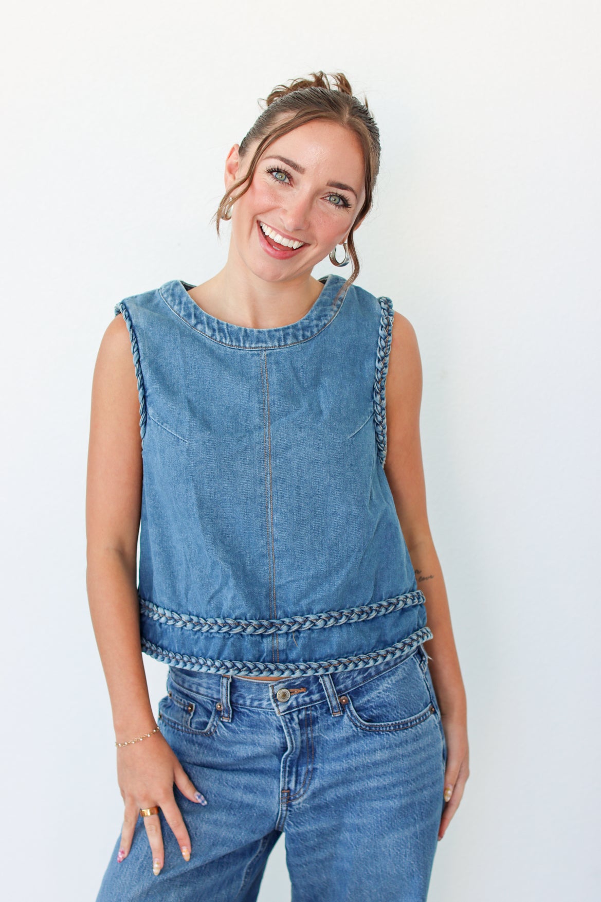 A smiling woman wearing a sleeveless blue denim top with decorative braided trim and matching blue jeans, standing against a plain white background.