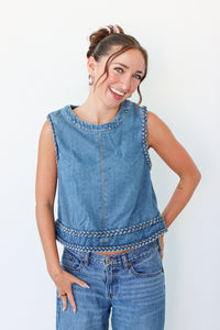 A woman modeling a sleeveless blue denim vest with decorative braided trim along the neckline and hem, paired with blue jeans against a plain white background.