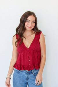 A smiling woman with long brown hair wearing a sleeveless deep red ruffled top with front buttons and blue jeans, standing against a plain white 
