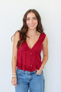 A smiling woman with long brown hair wearing a sleeveless deep red ruffled top with front buttons and blue jeans, standing against a plain white background.