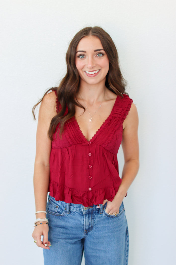 A smiling woman with long brown hair wearing a sleeveless deep red ruffled top with front buttons and blue jeans, standing against a plain white background.