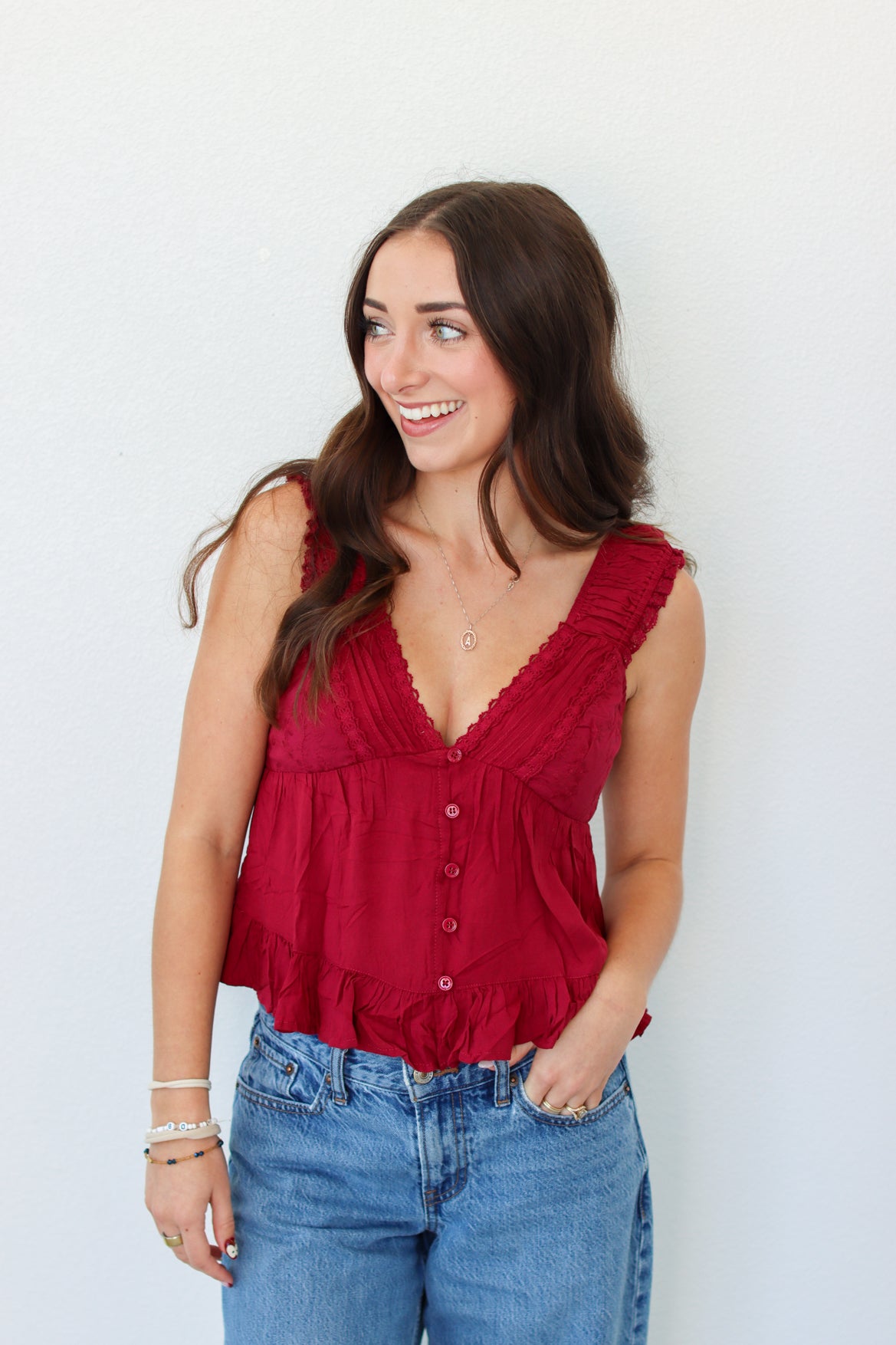 A woman wearing a sleeveless deep red ruffled top with a button front and blue jeans, standing against a plain white wall.