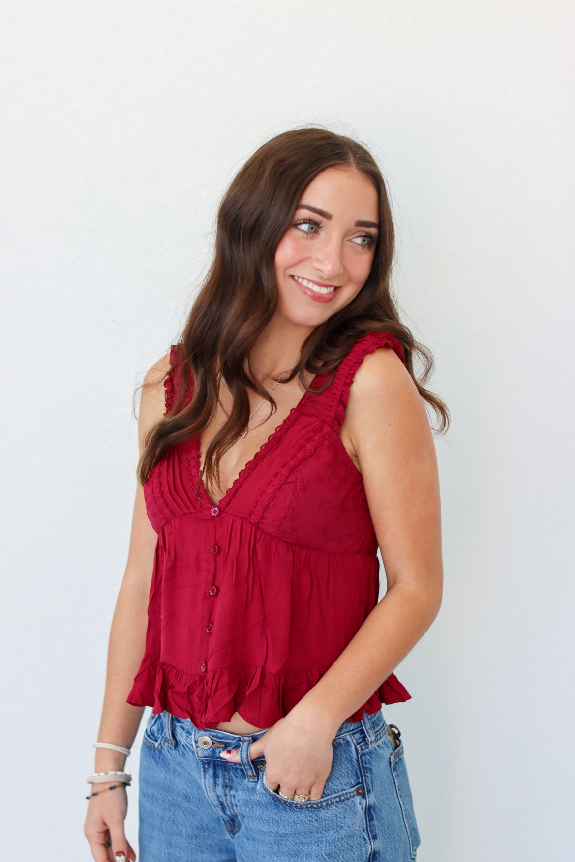 A woman with long brown hair wearing a sleeveless red ruffled top and blue jeans, standing against a plain white wall.