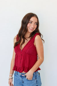 A woman with long brown hair wearing a sleeveless red ruffled top and blue jeans, standing against a plain white wall.