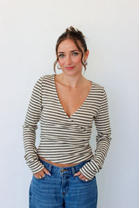 A woman wearing a beige and gray striped long-sleeve wrap top with blue jeans, standing against a plain white wall.