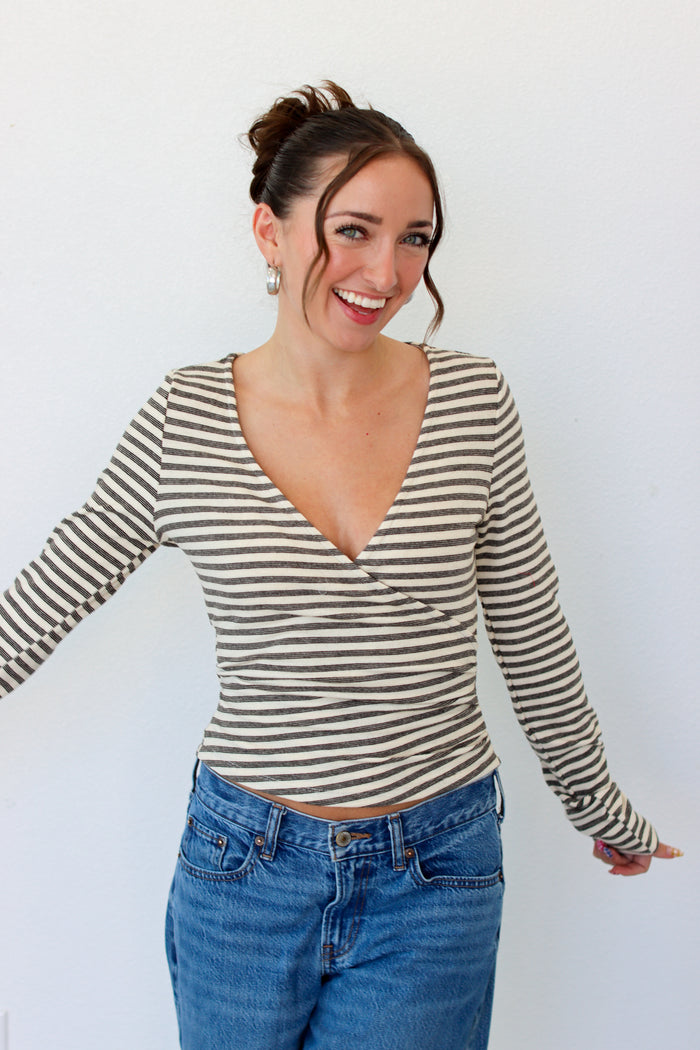 A woman wearing a beige and gray striped long-sleeve wrap top with blue jeans, standing against a plain white wall.