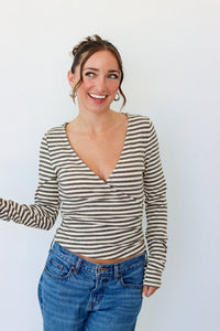 A woman wearing a beige and gray striped long-sleeve wrap top with blue jeans, standing against a plain white wall.