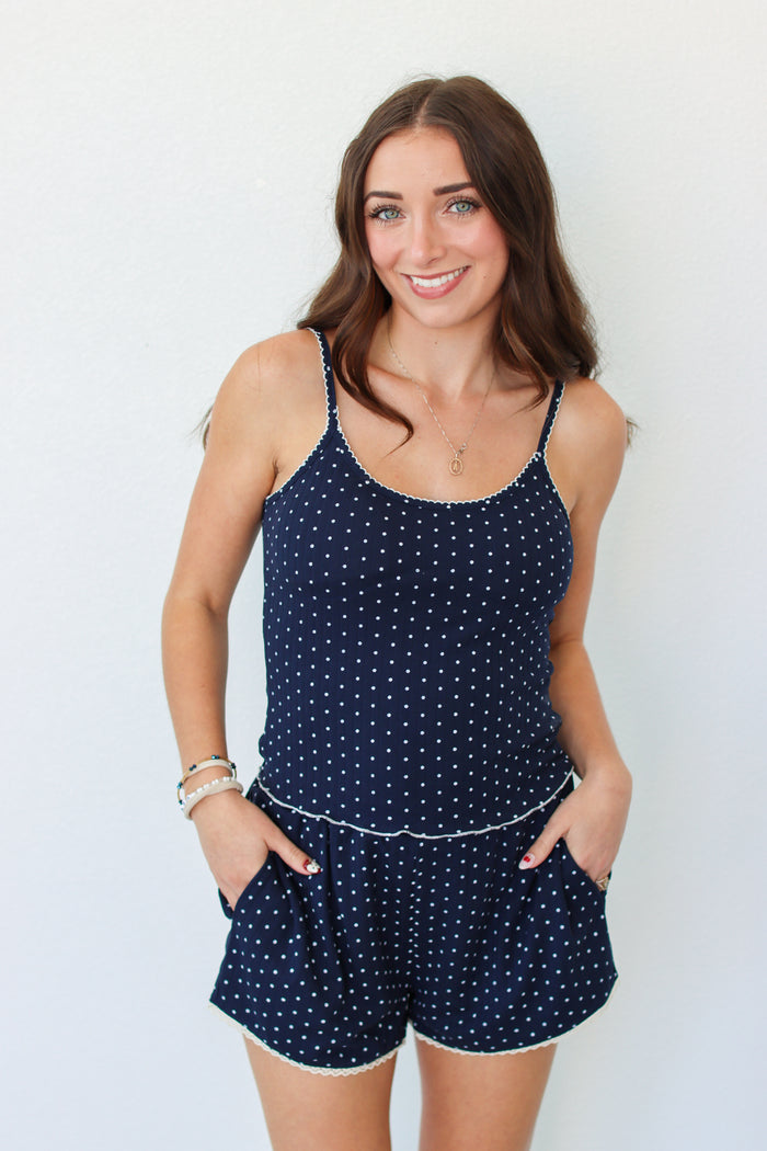 A woman modeling a navy blue tank and shorts set with white polka dots and white trim, standing against a plain white background.