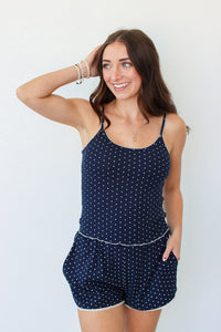 A woman modeling a navy blue tank and shorts set with white polka dots and white trim, standing against a plain white background.